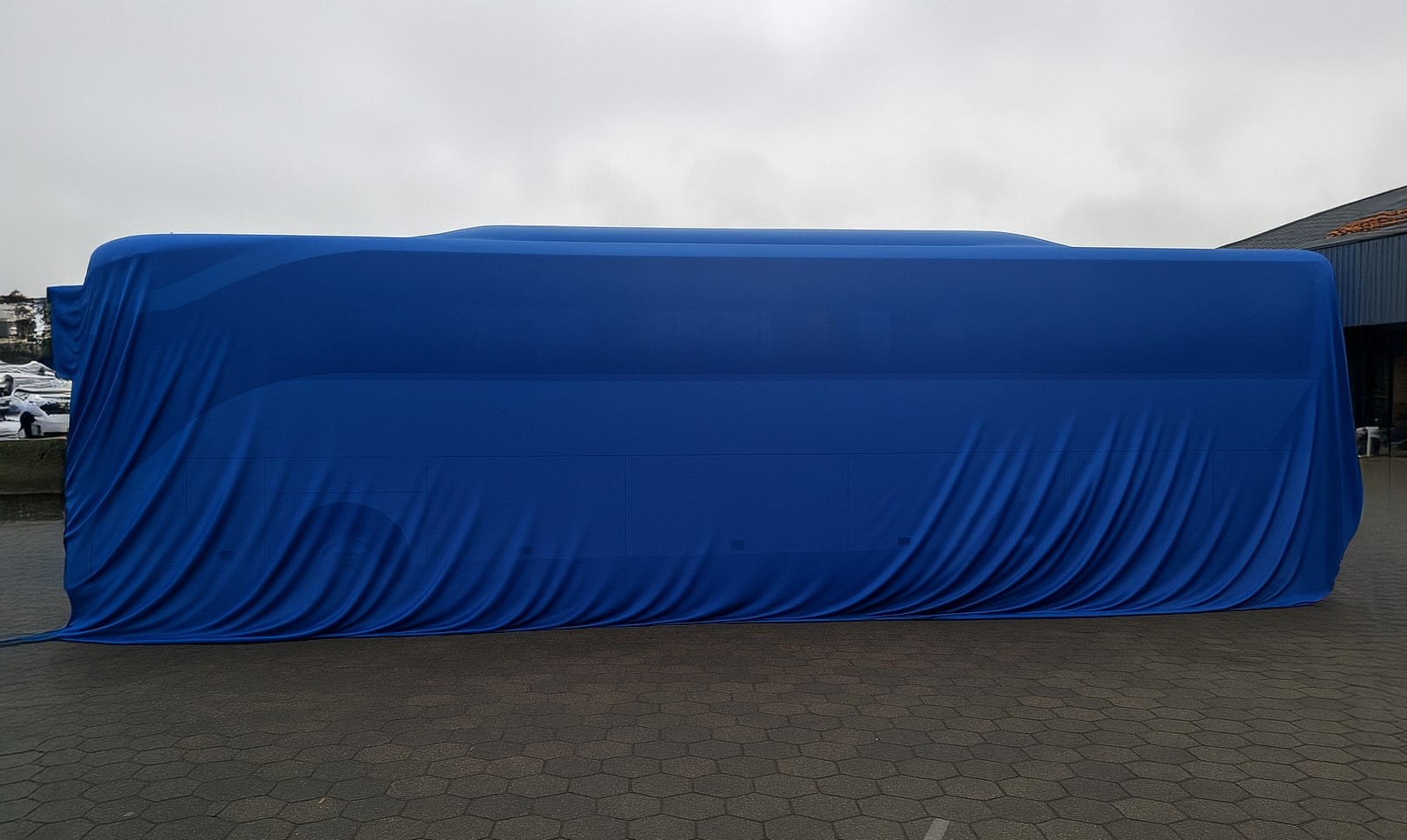 Een touringcar van Pouw Vervoer volledig bedekt met een strakblauw onthullingsdoek, klaar voor presentatie op een parkeerterrein met grijze bestrating en een bewolkte lucht op de achtergrond.